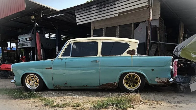 Ford Anglia 105E in blue and cream color, rust covered panels and gold aftermarket wheels.